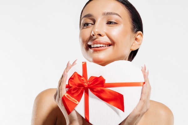 Smiling woman holding a heart-shaped gift box with a red ribbon. Dr. Pen Beauty Gifts are perfect for celebrating self-care