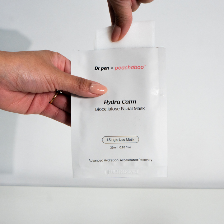 Close-up of a hand holding a Dr. Pen x Peachaboo Hydra Calm Biocellulose Facial Mask sachet with the mask partially visible inside the packet.