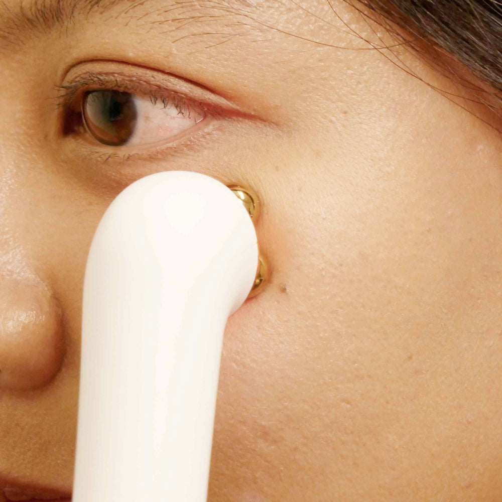 A woman gently cleanses her face with a white wand, showcasing the 3-in-1 Luxe Microcurrent Under Eye Massager.